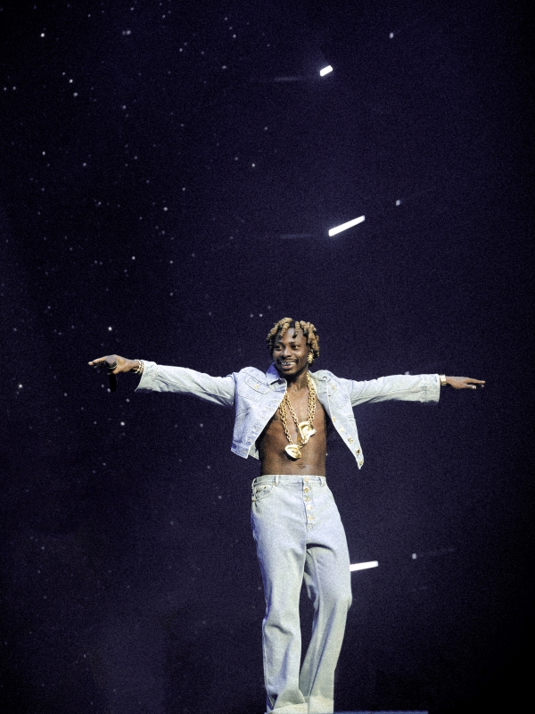 Asake in a white outfit with arms outstretched, performing under a single dramatic spotlight on a dark stage. — concert photography London by Lamss Media